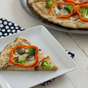 red pepper rings, broccoli, and mushrooms top a small pizza slice on a white plate on a black and white napkin