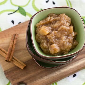 Chai-spied pear applesauce in a bowl on top of a stack of bowls next to two cinnamon sticks on a cutting board