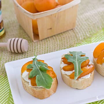 Apricot crostinis on a white plate next to a basket of ripe peaches