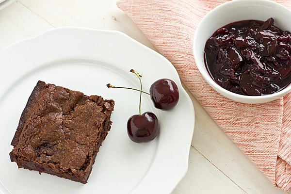 A bowl of cherry sauce sits next to a fresh-baked brownie on a china plate with fresh cherries beside it.