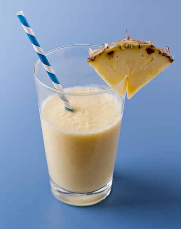 Frothy pineapple smoothie in a clear glass with a blue and white stripe straw sticking out and a pineapple wedge on the glass lip.