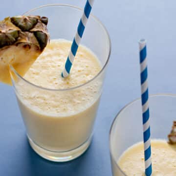 Frothy pineapple smoothies in glasses on a blue surface with blue and white straws