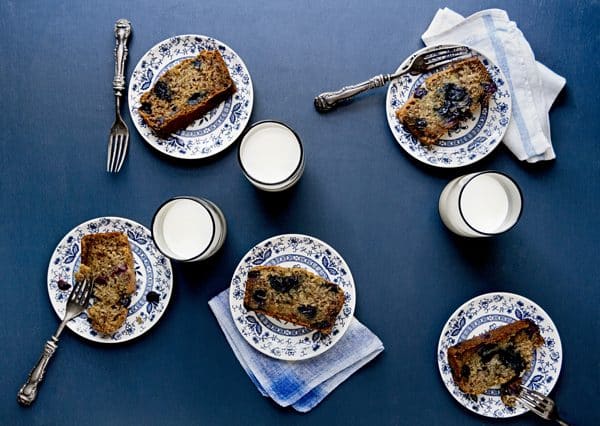 glasses of milk and small blue and white plates of blueberry bread sit on a dark blue table