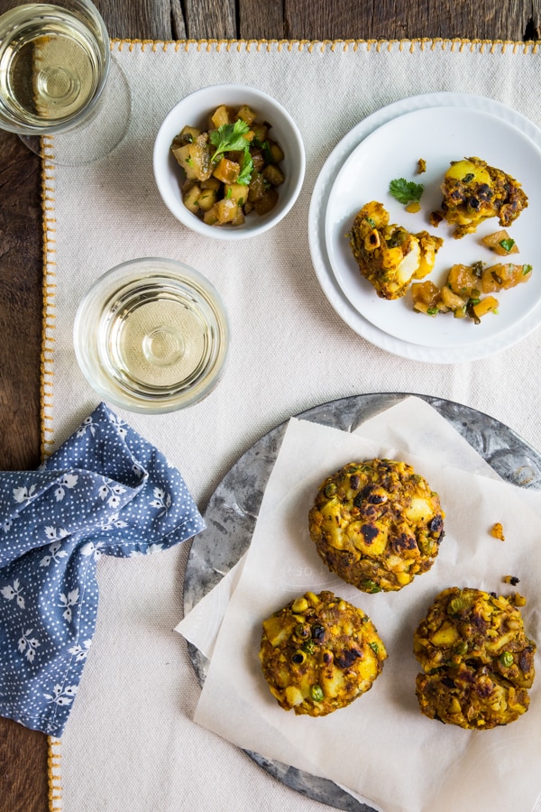 Two golden-brown samosa cakes on parchment paper next to a fork and a bowl of apple chutney
