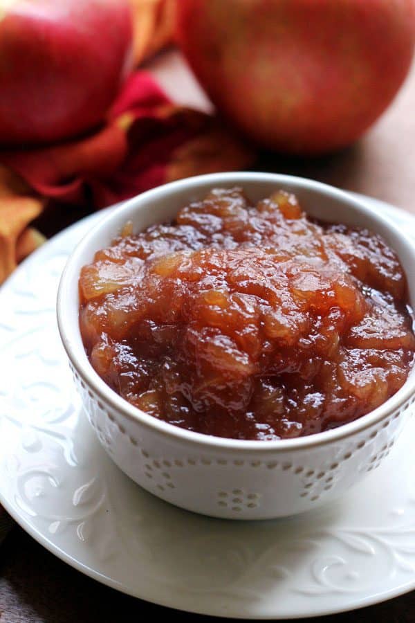 A white bowl of thick, deep brown homemade apple butter with whole apples in the background