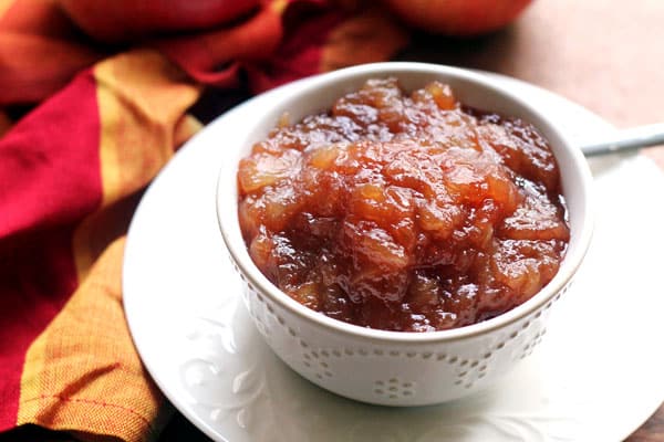 A bowl of finished homemade apple butter on a plate with a spoon and fresh apples nearby
