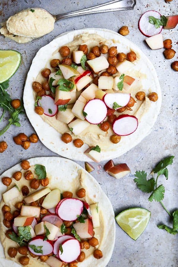 Two open-faced spiced chickpea tacos with hummus, roasted chickpeas, diced apple, and sliced radish on a metal tray