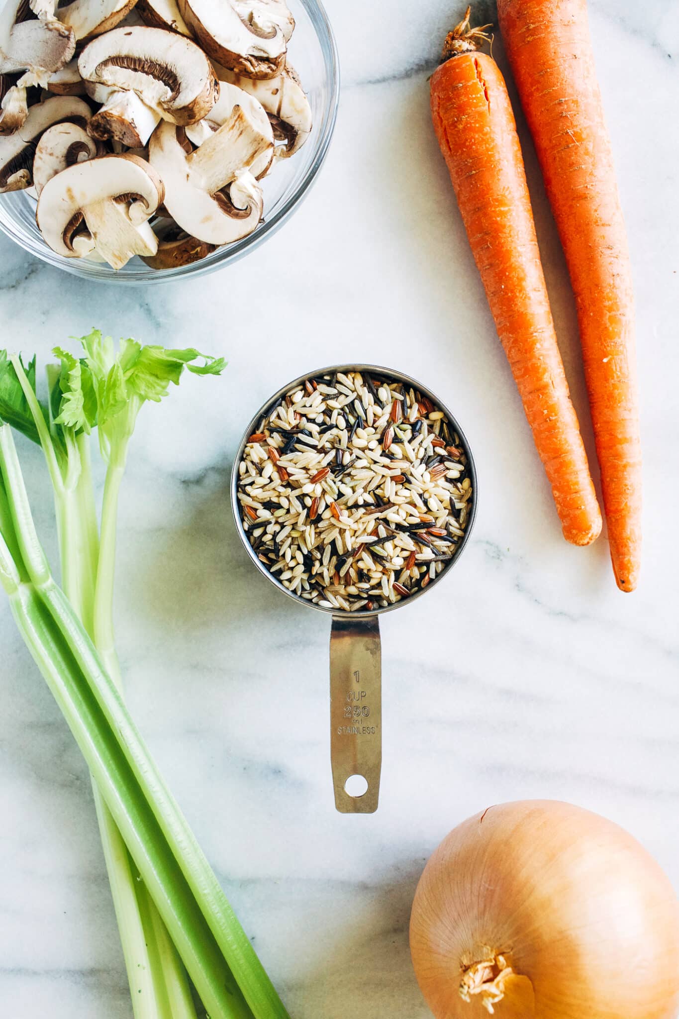 ingredients for making Creamy Wild Rice and Mushroom Casserole—bowl of sliced mushrooms, carrots, celery stalk, onion, and 1 cup measure full of wild rice