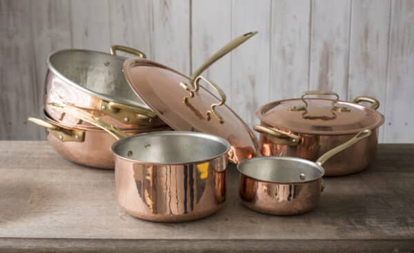 A set of copper cookware that include a small and medium-sized saucepan, a large saucepan, pot, and cover stacked on top of the other, and a covered small pot, all placed on a wooden countertop.