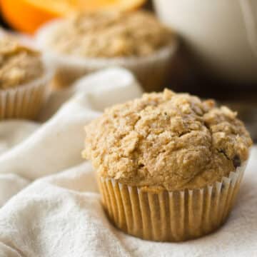 An orange spice muffin sits on a napkin in the foreground with other muffins and an orange out of focus behind