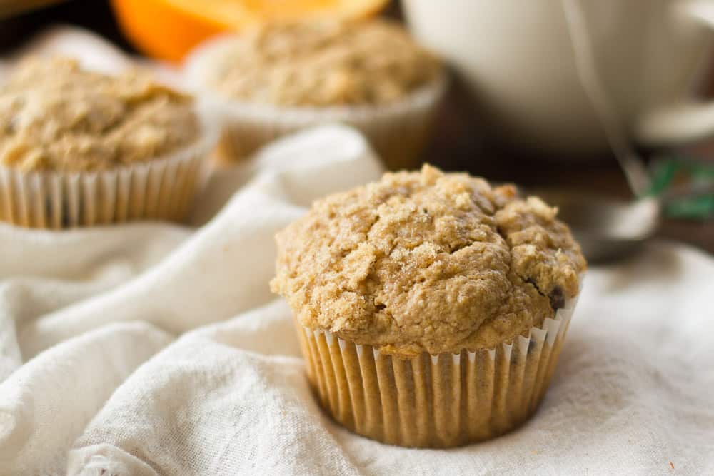 An orange spice muffin sits on a napkin in the foreground with other muffins and an orange out of focus behind