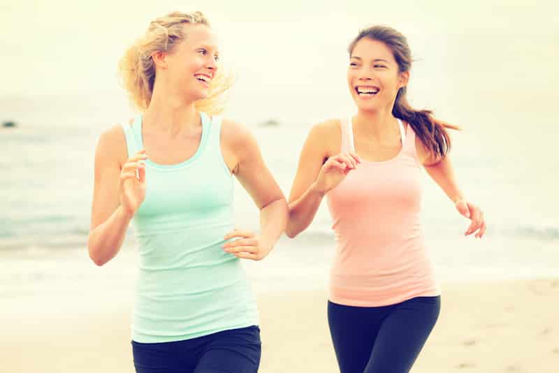 Women running exercising jogging happy on beach training as part of healthy lifestyle. Two fit female runners talking happy and smiling during workout. Multiracial Asian and Caucasian woman.
