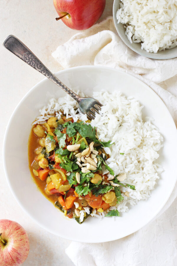 Bowl of vegetarian apple curry with chickpeas, kale, and cashews next to a bowl of basmati rice and fresh apples