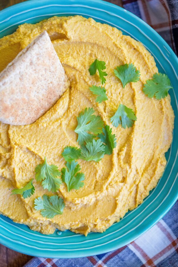 Pumpkin hummus in a teal bowl topped with cilantro, with a pita wedge dipped in