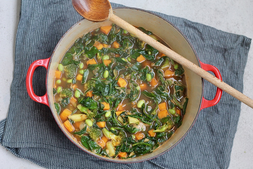 A large red soup pot is filled with the makings for winter green soup. Spinach, sweet potatoes, edamame, and broccoli make up the pot's contents and a wooden spoon rests on the lip of the pot.