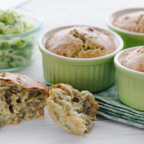 zucchini bread in mint green ramekins on a white counter behind a split zucchini bread muffin in front