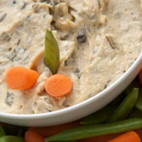 Artichoke hummus in a bowl surrounded by carrots and pea pods