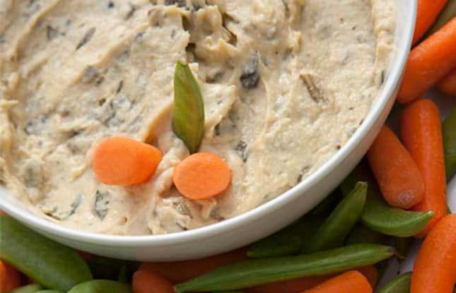 Artichoke hummus in a bowl surrounded by carrots and pea pods