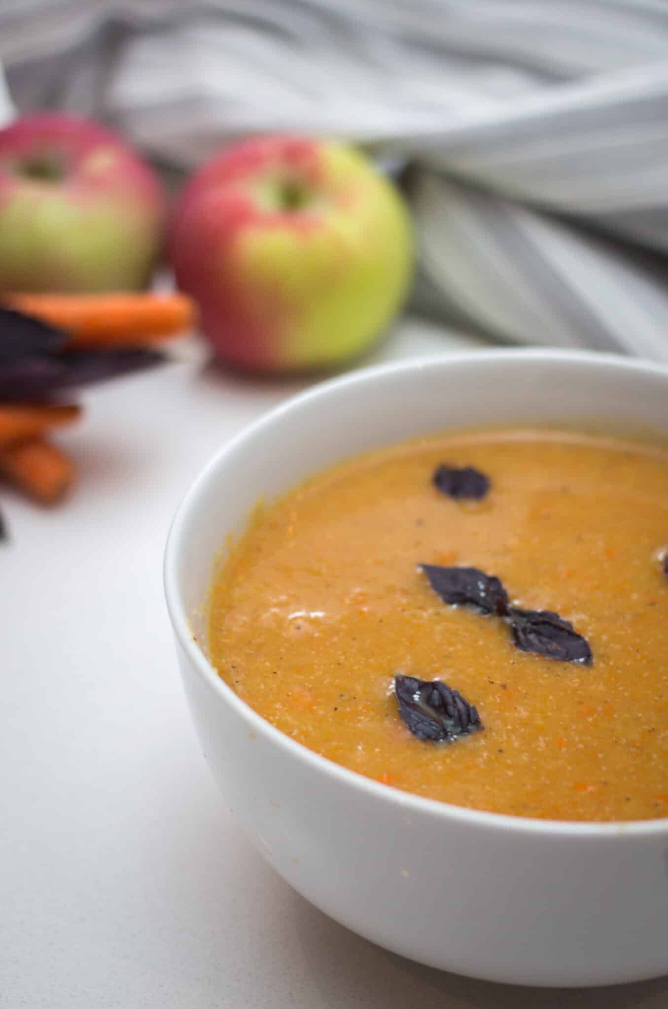 Vertical shot of a white bowl of apple and carrot soup topped with purple basil, with fresh carrots and apples in the background
