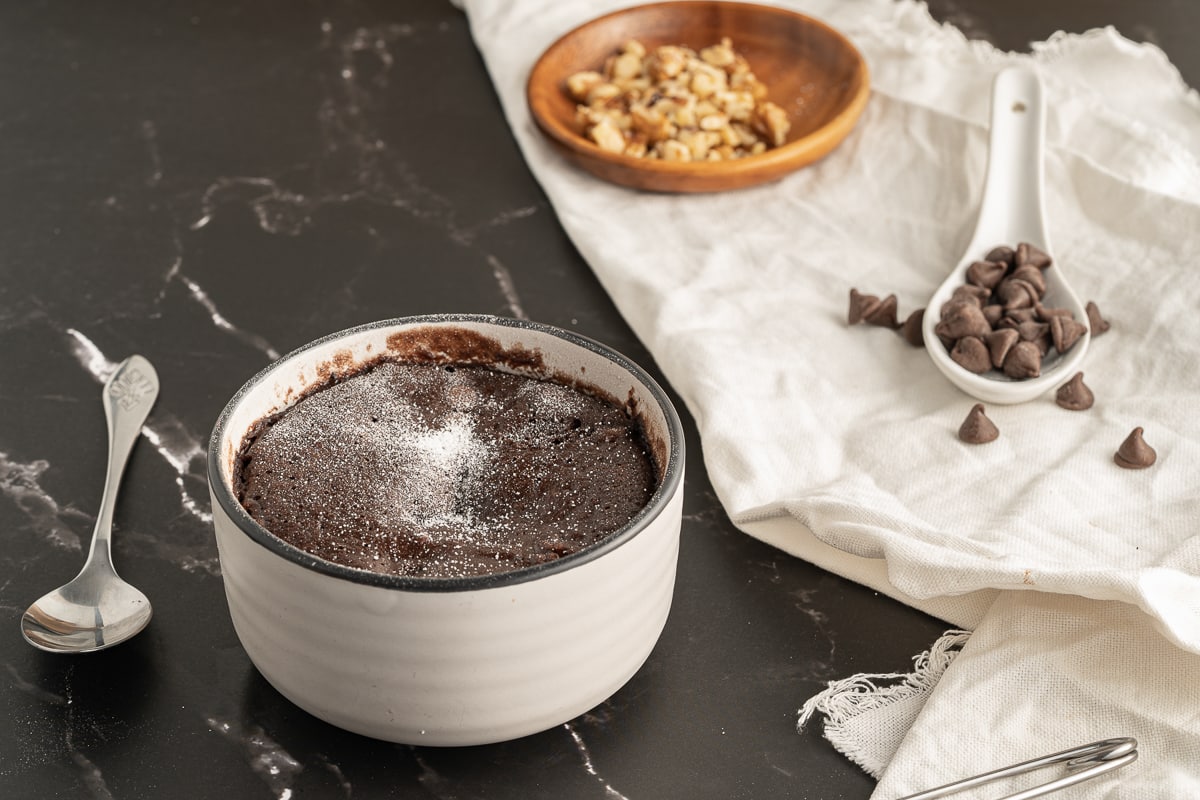 brownie mug cake on table with spoon, toppings, and cloth