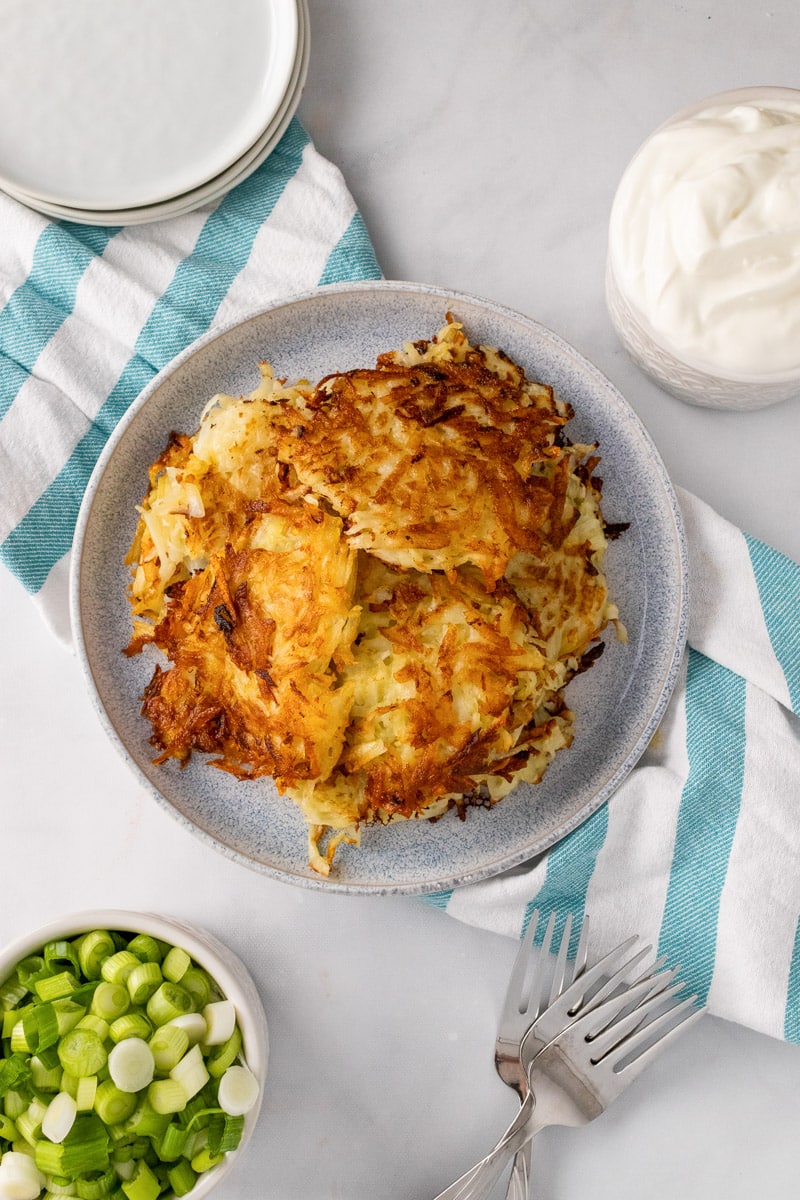 top view pile of potato pancakes on gray plate next small bowls of green onions and sour cream