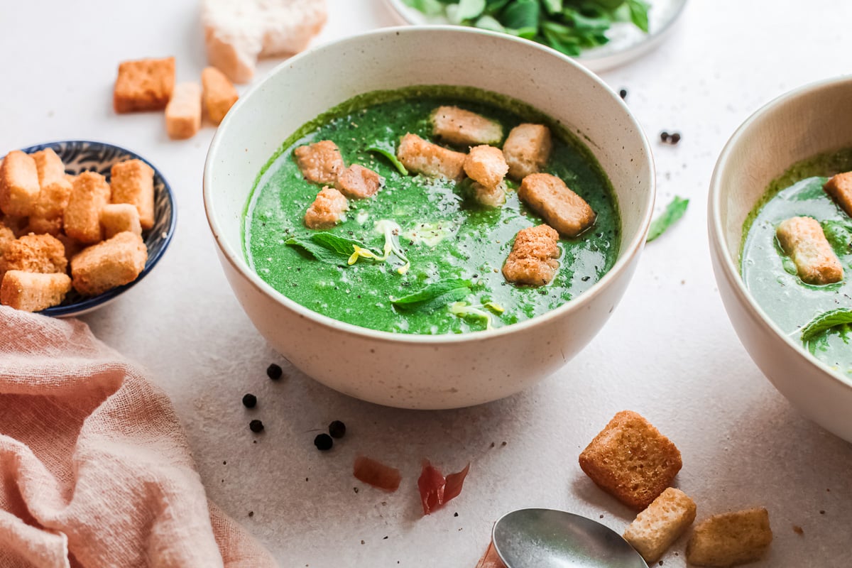 two bowls of vegan spinach soup next to small bowl of croutons and pink dish towel