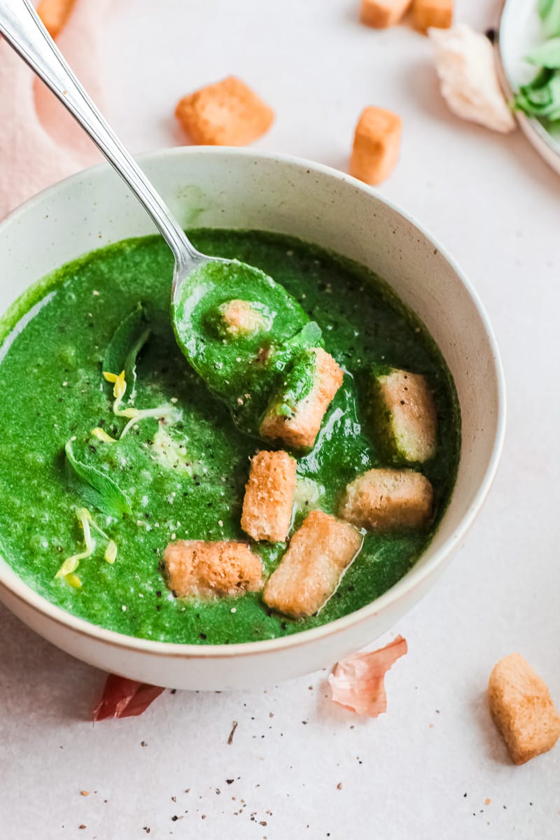 close up bowl of bright green vegan spinach soup with spoon and croutons