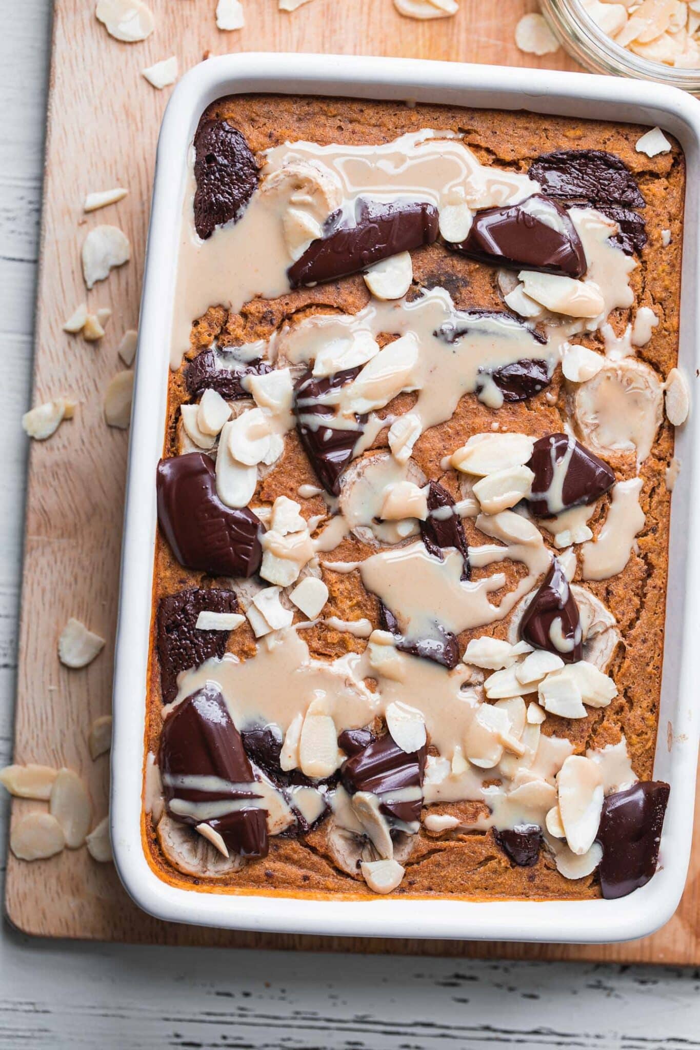Baked pumpkin oatmeal with dark chocolate and sliced almonds in a white baking dish on a wooden board