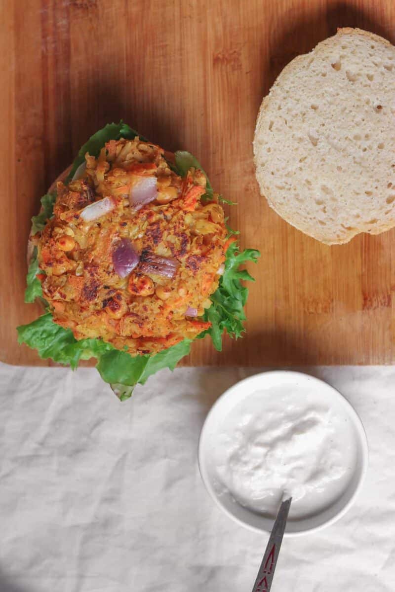 Vegan chickpea fritter burger resting on a table with a bowl of soy yoghurt nearby