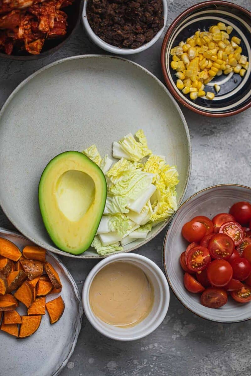 Bowl with avocado and lettuce surrounded by jackfruit, tomatoes, sweet potatoes, tahini, sweetcorn, and black beans