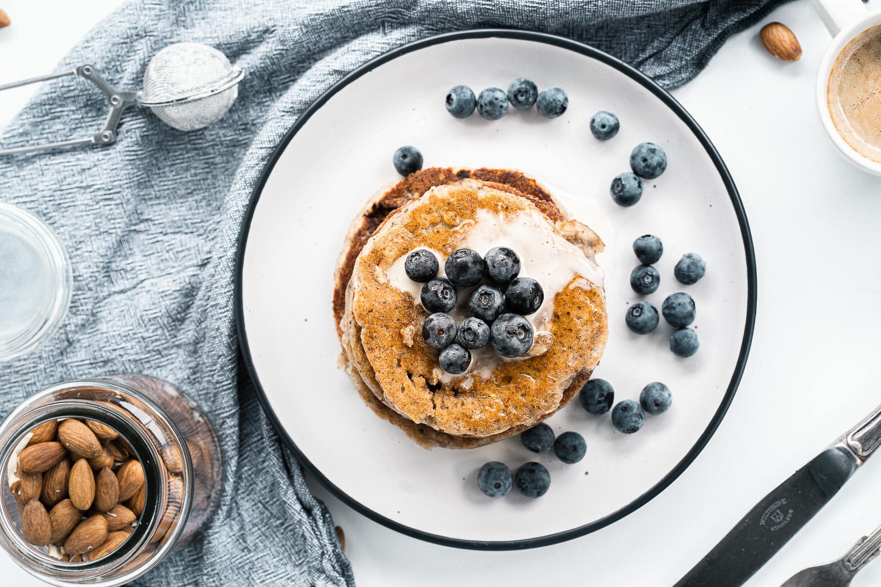 top view of stack of simple vegan pancakes with blueberries on a white plate with blue dishtowel and jar of almonds