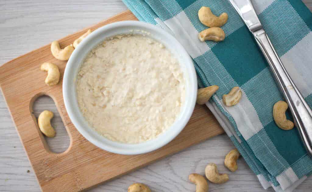 ramekin of homemade vegan cream cheese on a wooden board with butter knife, cashews, and blue checked dish towel