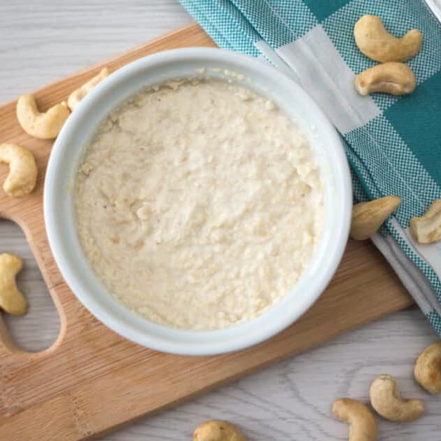 close up ramekin of homemade vegan cream cheese surrounded by cashews with wooden board and dish towel