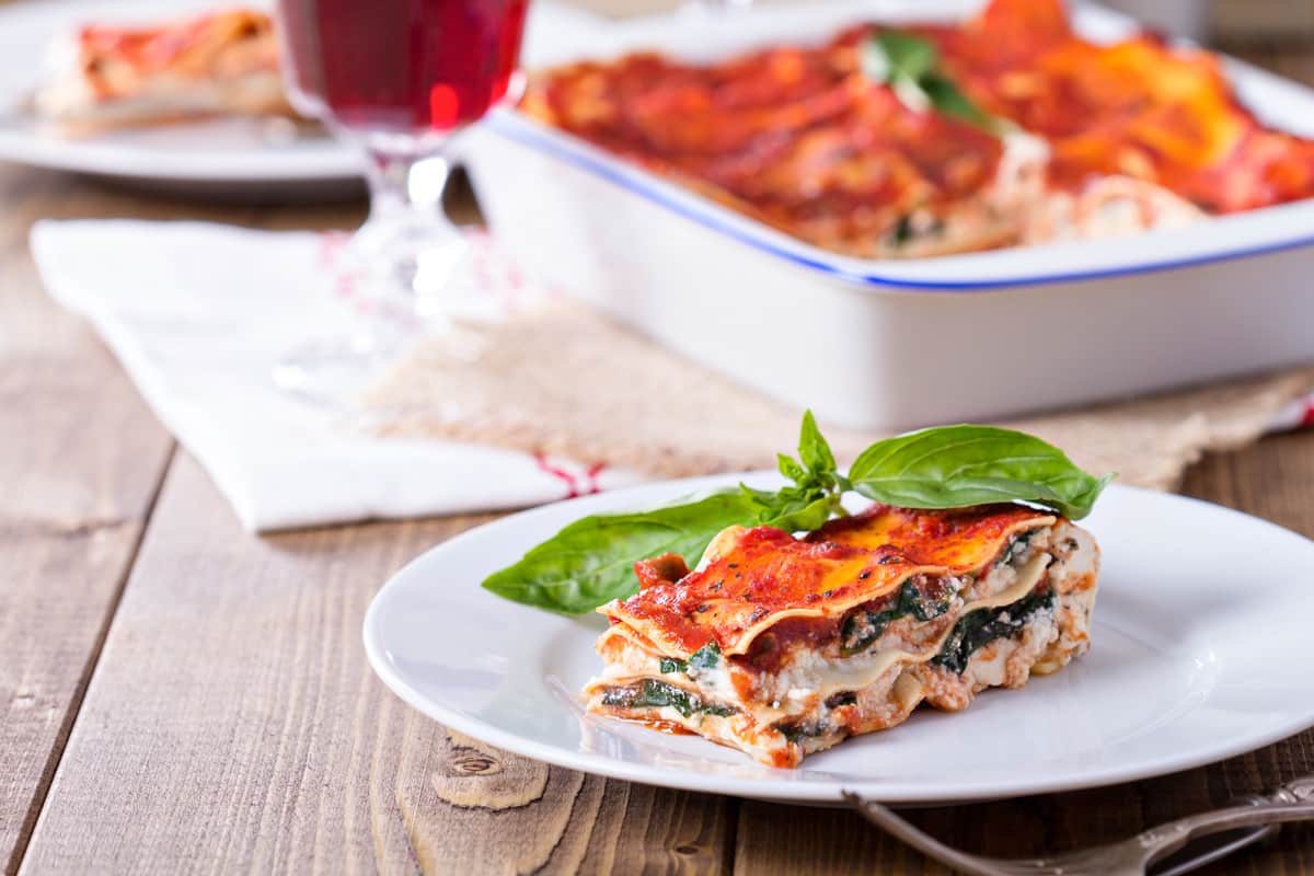 far out wide view of slice of best vegan lasagna on white plate with casserole dish and table settings in background on wooden table