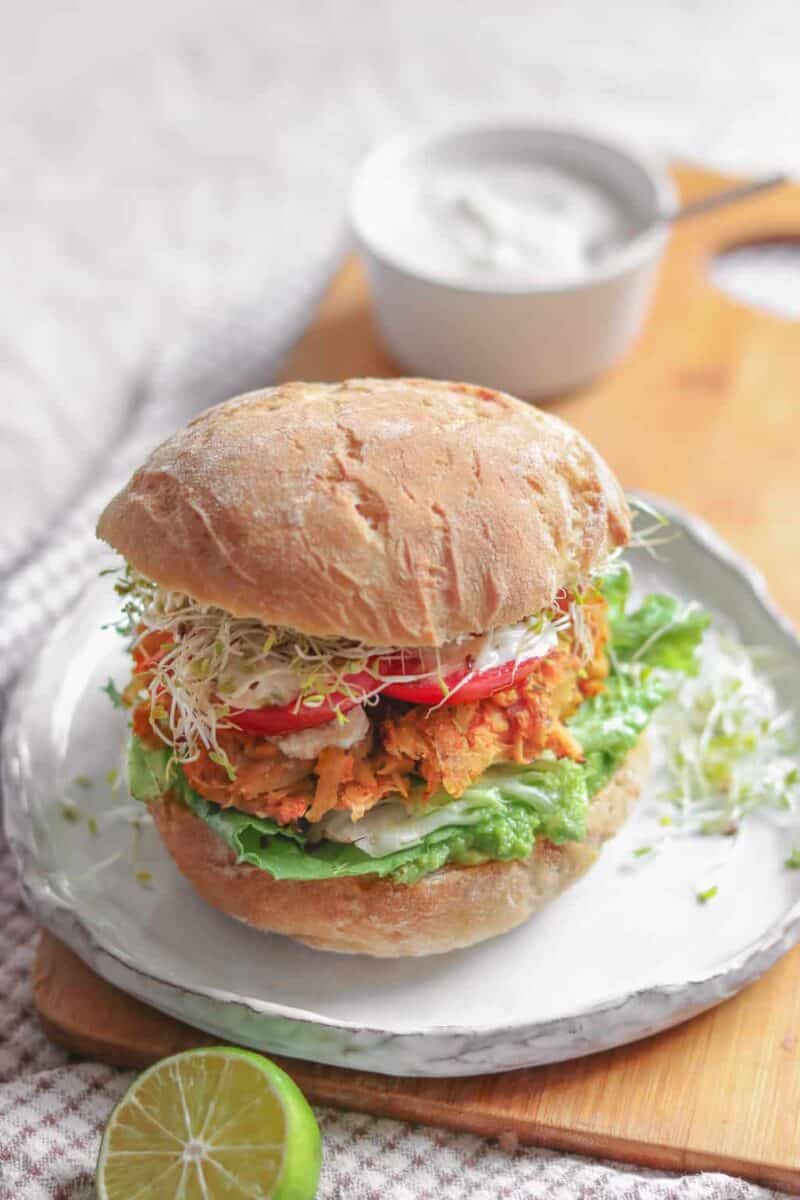 Vegan sandwich with chickpea fritters, avocado, alfalfa sprouts, tomatoes, mixed greens and soy yoghurt on a white plate