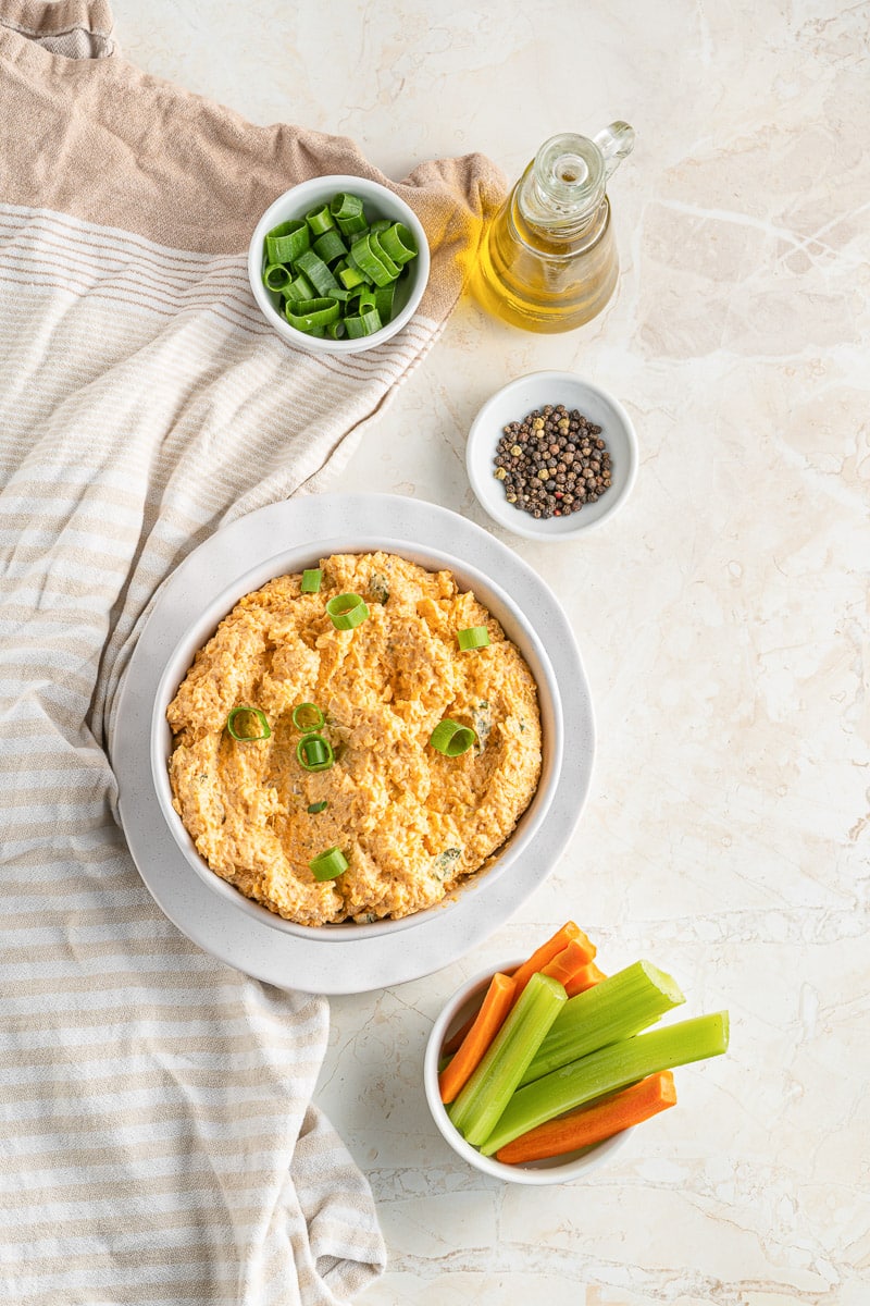 buffalo cauliflower dip in a bowl next to small bowls of ingredients and celery sticks