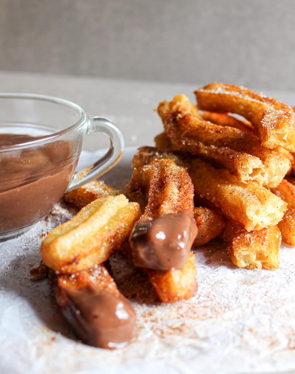 stack of homemade churros with a few ends dipped in chocolate next to a bowl of melted chocolate