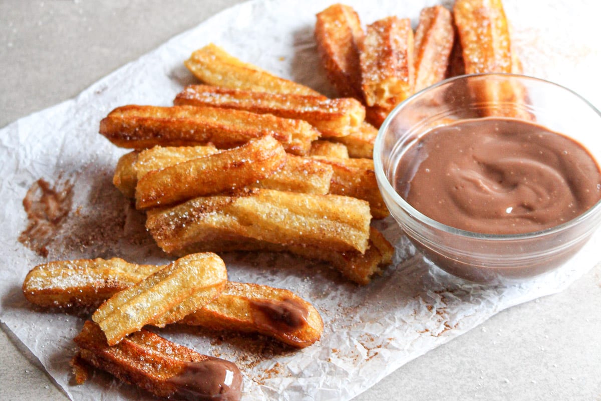 pile of homemade churros on wax paper next to a bowl of melted chocolate