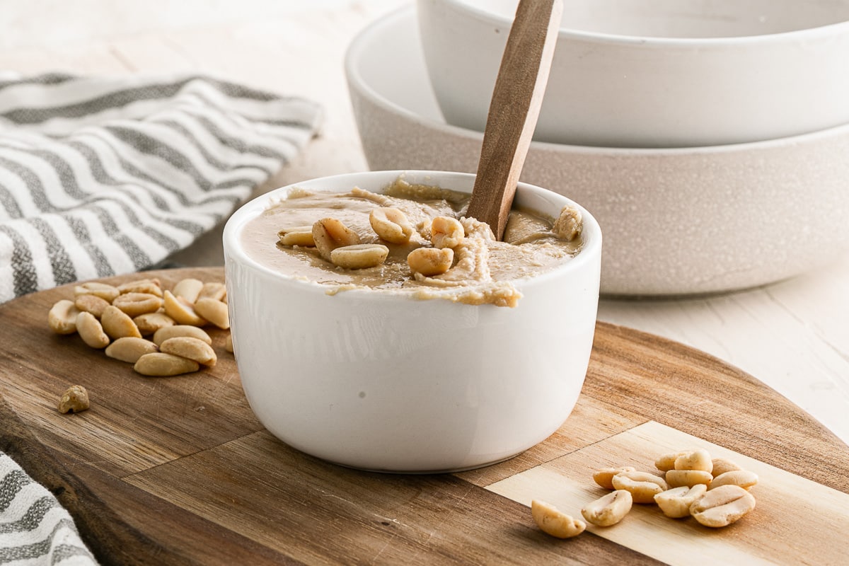 small white bowl of 5-minute homemade peanut butter with wooden spoon on wooden board with dishtowel behind and sprinkled with peanuts