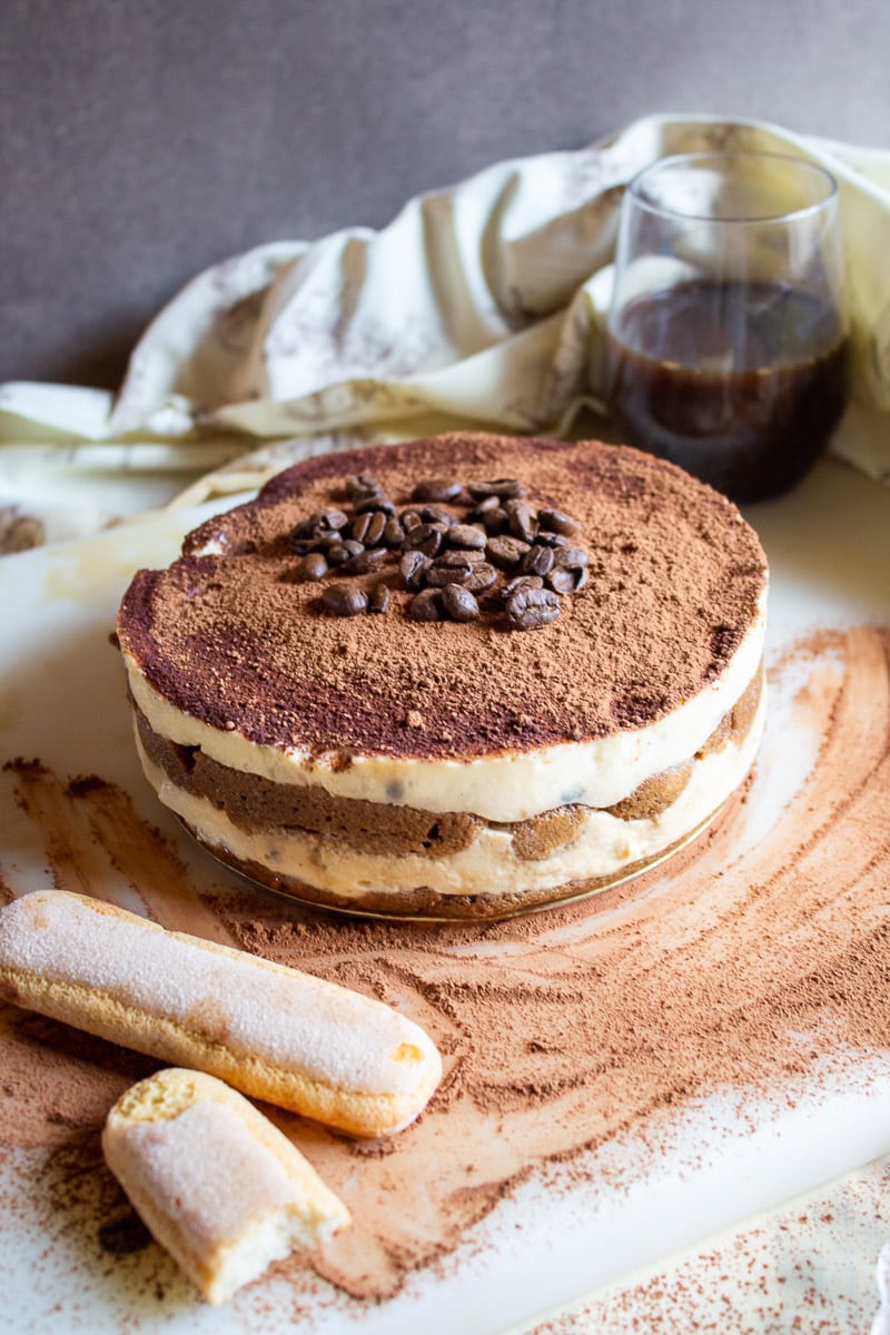 entire best homemade tiramisu next to two ladyfingers on cocoa-sprinkled countertop