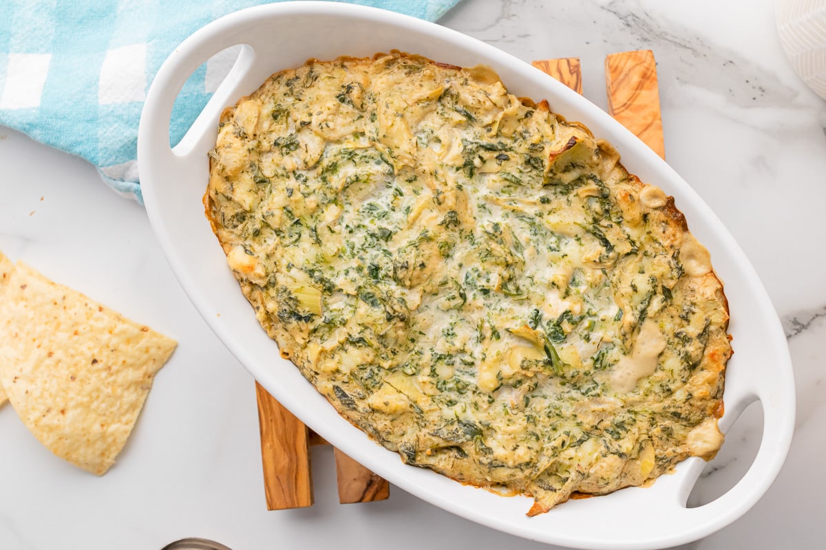 top view of baked spinach artichoke dip without mayo in white casserole dish with chips and blue dishtowel at the side