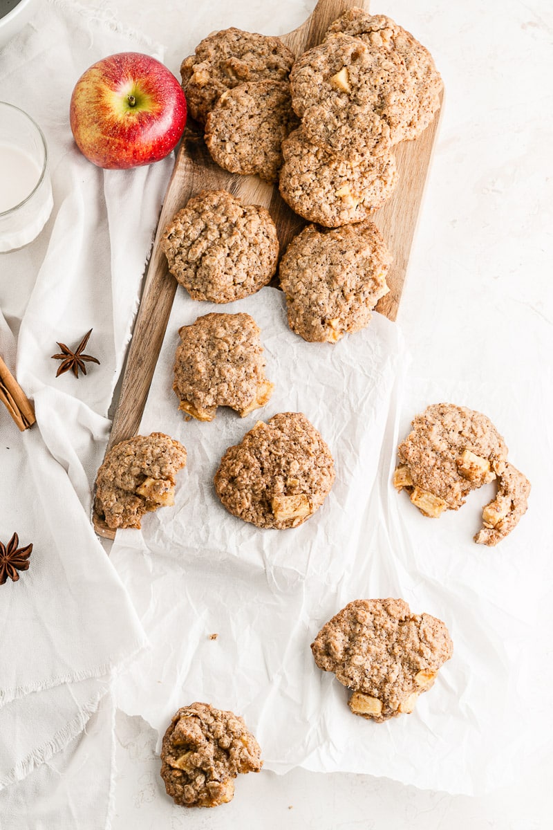 vegan apple oatmeal cookies scattered over wooden serving board with apple, cloves, and cinnamon stick