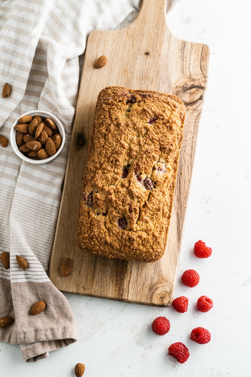 loaf of raspberry bread on a wooden bread board next to almonds, raspberries, and dish towel