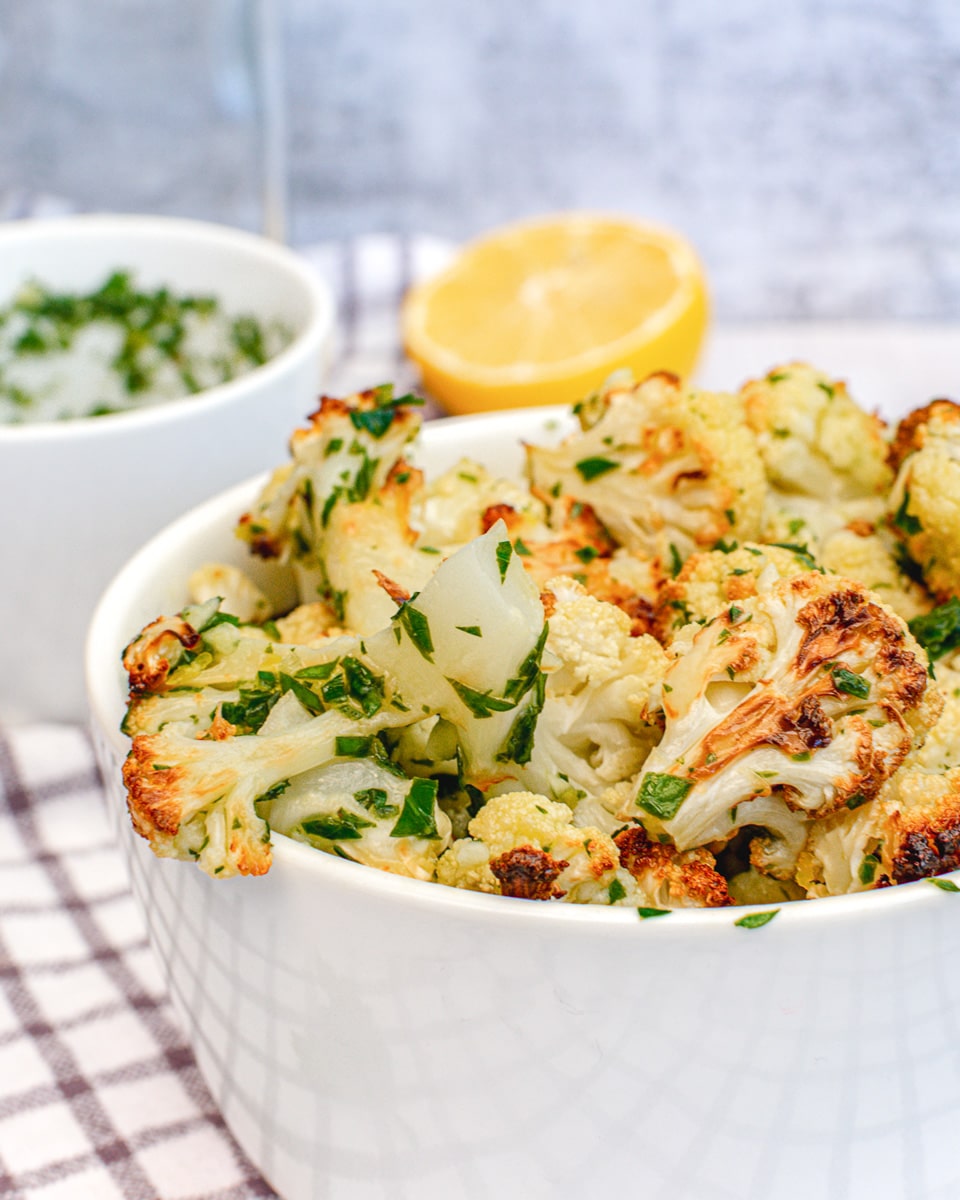 close up roasted cauliflower with lemon, garlic, and herbs in white bowl on table