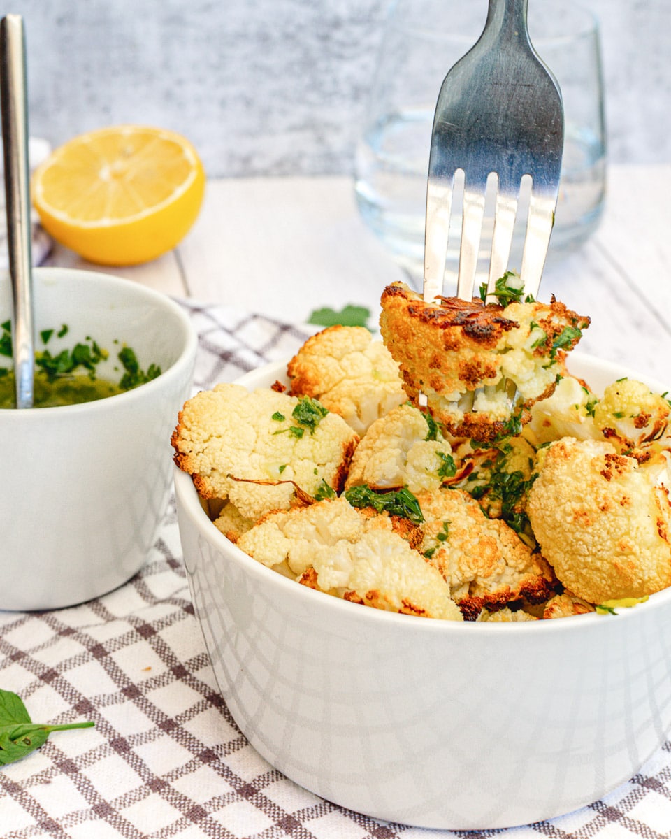 fork picking up roasted cauliflower with lemon, garlic, and herbs out of white bowl on table