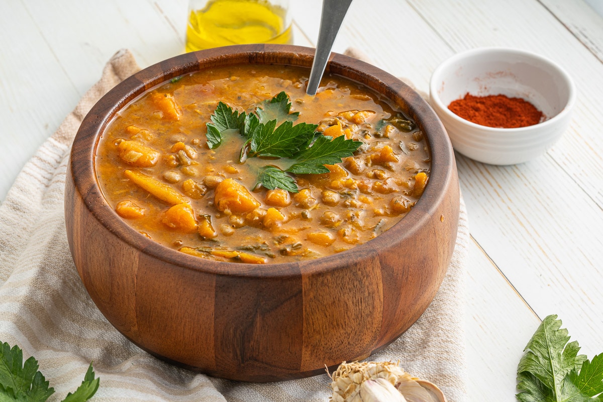 side view of wooden bowl full of one-pot sweet potato lentil stew with spoon next to small bowls of spices and oil