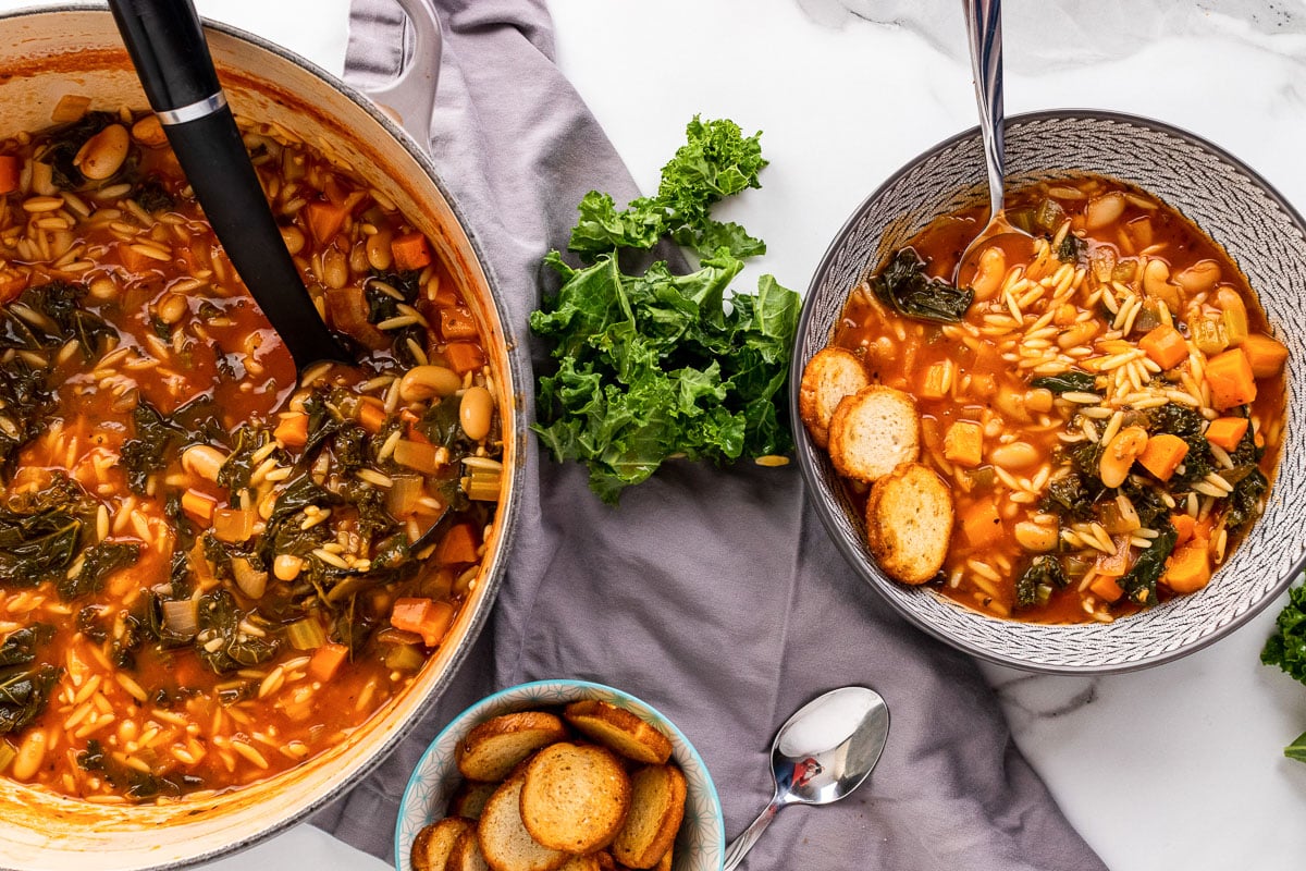 pot of tuscan kale soup with black ladle next to bowl of soup with silver spoon and croutons