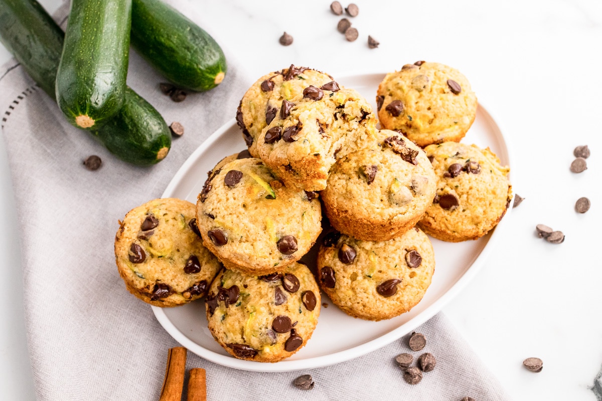 white plate stacked with zucchini chocolate chip muffins on white tablecloth scattered with chocolate chips, zucchinis, and cinnamon sticks