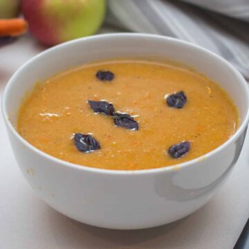 Overhead close-up of creamy apple and carrot soup in a white bowl, garnished with purple basil leaves