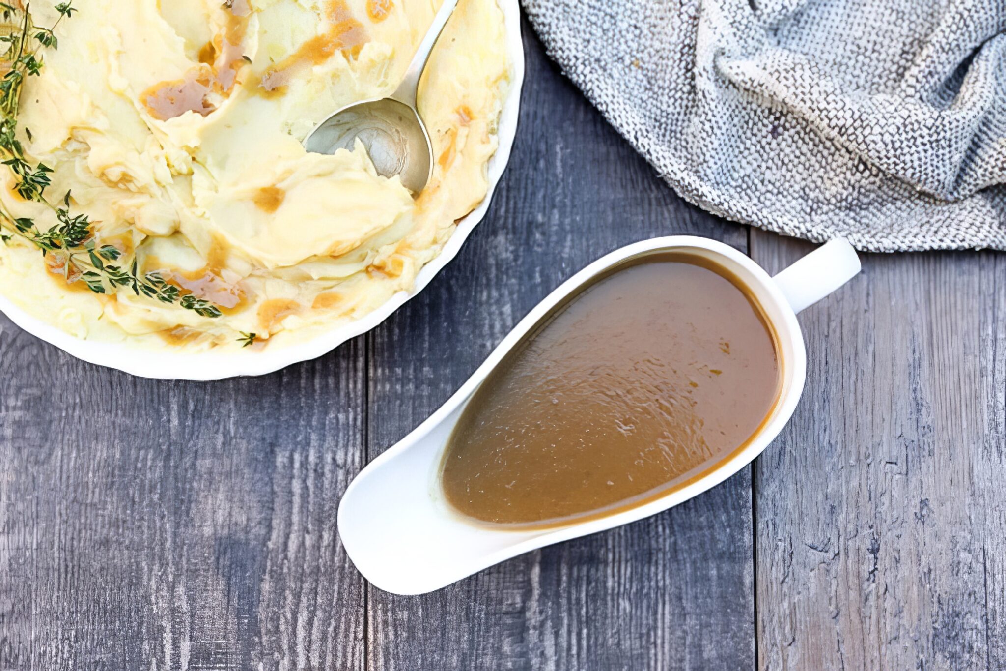 Wide overhead shot of apple cider gravy in a gravy boat beside herb-topped mashed potatoes on a rustic wood table